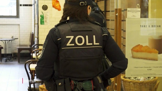 Eine Mitarbeiterin des Zolls in dunkelblauer Uniform von hinten, auf dem Rücken ihrer Weste steht in Großbuchstaben "ZOLL". © Screenshot 