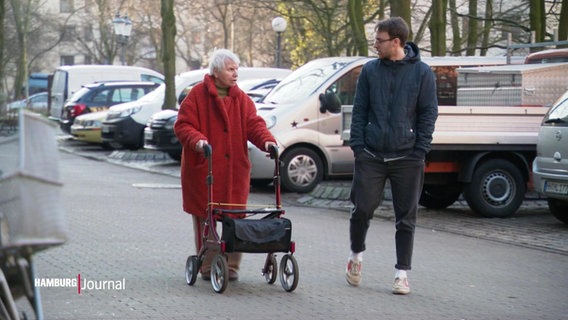 Eine alte Dame mit Rollator und ein junger Mann gehen zusammen eine Straße entlang. © Screenshot 