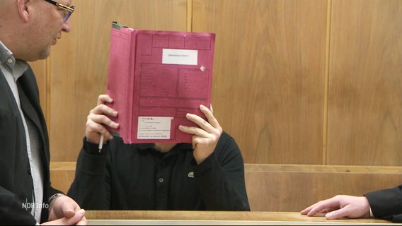 Ein Mann schützt sein Gesicht im Gerichtssaal mit einer Akte. © Screenshot 