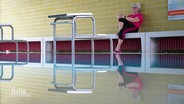 Petra Seipelt sitzt auf einer Bank im Hallenbad, im Vordergrund ist das Schwimmbecken mit Springblöcken zu sehen. © Screenshot 