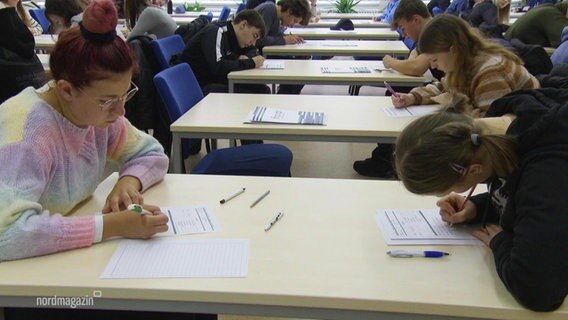 Mehrere junge Leute sitzen an verschiedenen Tischen und schreiben etwas auf. © Screenshot 