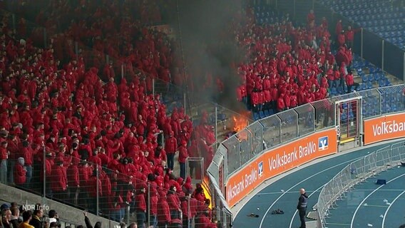Rotgekleidete Fans stehen im Qualm im Zuschauerbereich. © Screenshot 