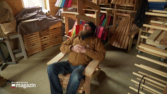 Ein Mann sitzt in einem hölzernen Liegestuhl in einer Werkstatt. © Screenshot 
