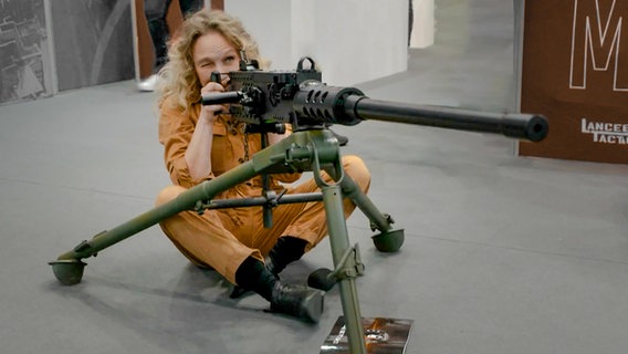 Reporterin Katja Kreml sitzend vor einer Waffe bei der Waffenmesse in Nürnberg. (extra 3 vom 23.03.2023 im Ersten) © NDR 