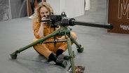 Reporterin Katja Kreml sitzend vor einer Waffe bei der Waffenmesse in Nürnberg. (extra 3 vom 23.03.2023 im Ersten) © NDR 
