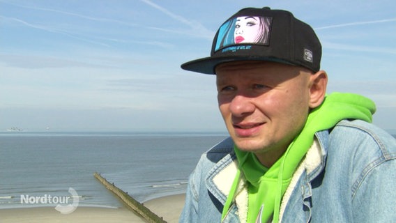 Der junge Mann Radek Krajewski im Interview. Er trägt ein Käppi und im Hintergrund ist ein Strandabschnitt zu sehen. © Screenshot 