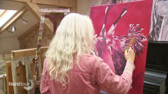 Eine Malerin malt ein rotes Bild mit Kranichen. Die Malerin ist von hinten zu sehen. © Screenshot 