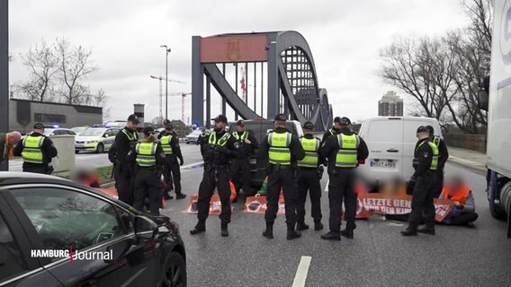 Mehrere Polizisten stehen um Klimaprotestierende, die auf einer Straße sitzen. © Screenshot 