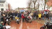 Viele Menschen stehen im Regen auf einem Platz. Vor ihnen stehen einige Mororräder. © Screenshot 