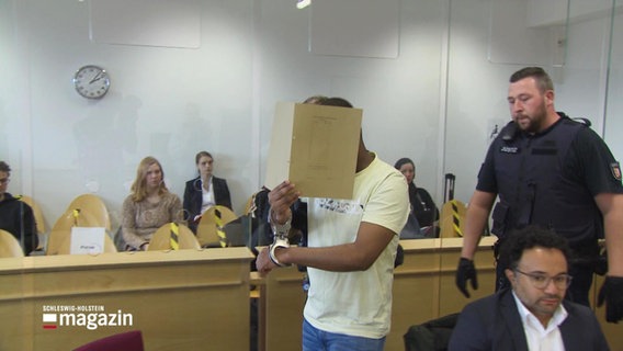 Ein Mann in einem Gerichtssaal verdeckt sein Gesicht mit einer Mappe. © Screenshot 