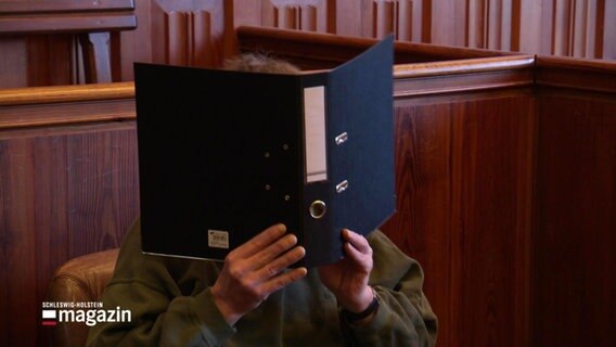 Eine Person in einem Gerichtssaal verdeckt ihr Gesicht mit einem Ordner. © Screenshot 