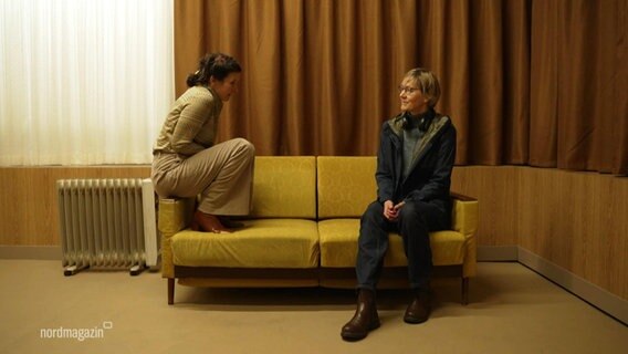 Zwei junge Frauen sitzen auf einer gelben Couch. Die Jüngere von beiden auf der linken Lehne. Sie sieht die zweite auf der rechten Seite des Möbelstück an. © Screenshot 