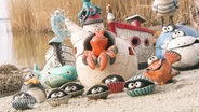 Mehrere bunt bemalte Keramik-Figuren mit maritimen Zügen sind auf einer Sandfläche an einem Ufer angeordnet. © Screenshot 