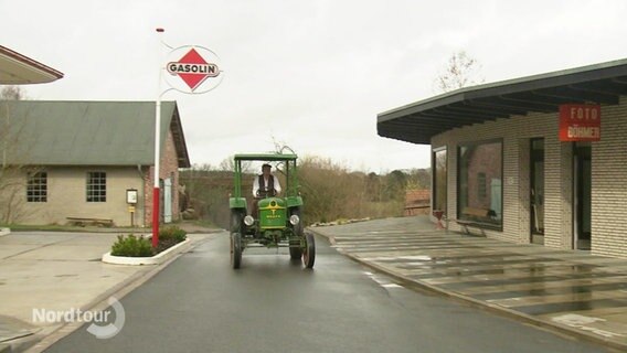 Ein Mann fährt einen Traktor-Oldtimer auf einer Straße die flankiert wird von einer älteren Tankstelle im Stil der 50er Jahre und einem älteren Geschäft für Radiogeräte. © Screenshot 