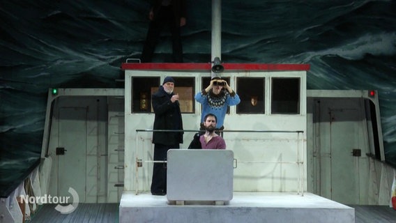 Auf einer Theaterbühne stehen mehrere Darstellende auf einem Schiffsdeck und schauen mit einem Fernglas in die stürmische Wellenfront. © Screenshot 