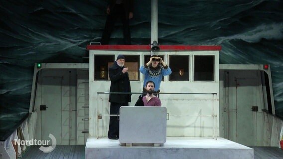Auf einer Theaterbühne stehen mehrere Darstellende auf einem Schiffsdeck und schauen mit einem Fernglas in die stürmische Wellenfront. © Screenshot 