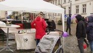 Auf einem verregneten Frühlingsfest stehen einige Menschen unter weißen Pavillion-Zelten. © Screenshot 