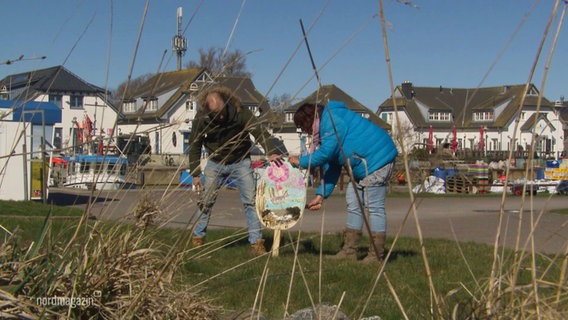 Zwei Personen stecken ein großes Schild in Osterei-Form in eine Grasfläche. © Screenshot 