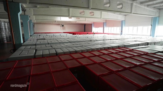 In einer größeren Halle lagern viele Kisten in mehreren Stapelreihen. © Screenshot 