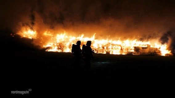 Zwei Feuerwehrleute stehen nachts vor einem Brand. Die Flammen schlagen in die Höhe. © Screenshot 