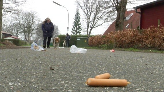 Auf einer Straße liegen zwei Würstchen, im Hintergrund geht eine Frau mit einem Hund an der Leine. © Screenshot 