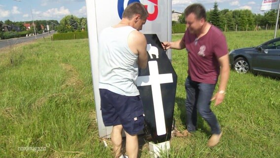 Zwei Beschäftigte stellen aus Protest vor einem Fahrzeugwerk einen kleinen Sarg auf. © Screenshot 