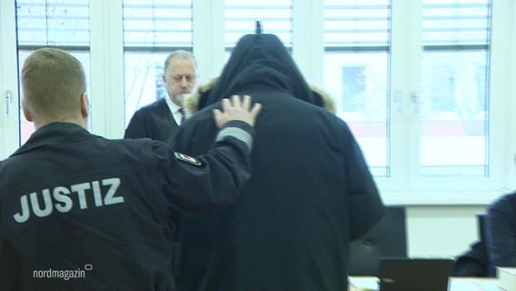 Ein Justizbeamter führt eine Person mit schwarzer Kapuzenjacke in einen Gerichtssaal. © Screenshot 