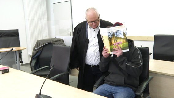 Vor Gericht hält sich eine Angeklagter ein Heft vors Gesicht, sein Anwalt steht neben ihm. © Screenshot 