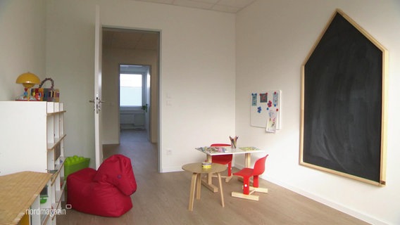 Blick in einen Raum mit geöffneter Tür in dem sich ein kleiner Kinder-Maltisch, Pferde-förmiges Sitzkissen und eine Tafel befinden. © Screenshot 