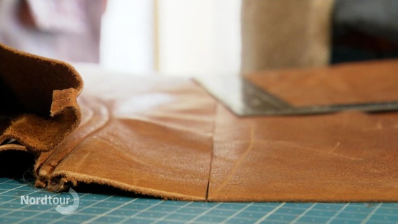Nahaufnahme: Auf einer Arbeitsfläche liegen ein Stück Leder und ein Lineal. © Screenshot 
