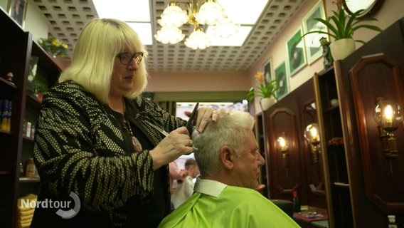 Eine Frau schneidet einem Mann die Haare in einem historisch anmutenden Friseursalon. © Screenshot 