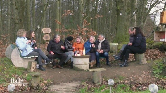 Einige Menschen sitzen auf Bänken an einem Waldrand und trinken Getränke. © Screenshot 