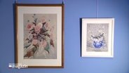 An einer blauen Wand hängen zwei unterschiedliche Blumen-Bilder. © Screenshot 