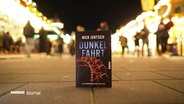 Auf einem Weg aus Pflastersteinen steht ein Buch mit dem titel "Dunkelfahrt", im Hintergrund: mehrere hell leuchtende Buden eines Jahrmarkts. © Screenshot 