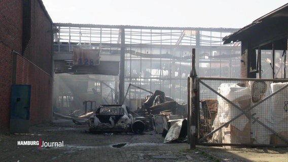 Auf einem Betriebsgelände steht ein abgebranntes Autowrack, im Hintergrund eine durch einen Brand zerstörte Lagerhalle. © Screenshot 