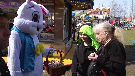Ein größeres Osterhasenmaskottchen hält einem Jungen und einer Frau auf einem Jahrmarkt einen Korb mit Ostereiern hin. © Screenshot 