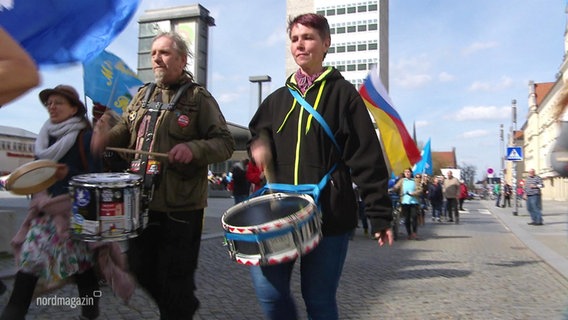 Demonstrierende gehen bei einem Ostermarsch mit Trommeln vor dem Bauch durch die Straßen. © Screenshot 
