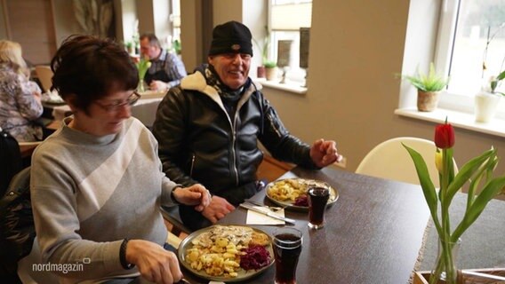 Eine Frau und ein Mann sitzen grinsend an einem Esstisch in einer Gaststätte, vor ihnen: jeweils Teller mit Schnitzel und Pilzsoße. © Screenshot 