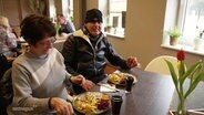 Eine Frau und ein Mann sitzen grinsend an einem Esstisch in einer Gaststätte, vor ihnen: jeweils Teller mit Schnitzel und Pilzsoße. © Screenshot 