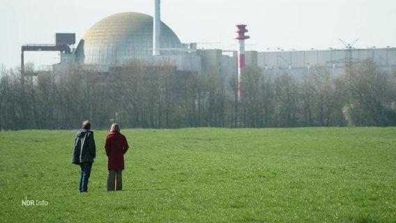 Zwei Menschen stehen auf einer grünen Wiese, im Hintergrund ist ein Atomkraftwerk zu sehen. © Screenshot 