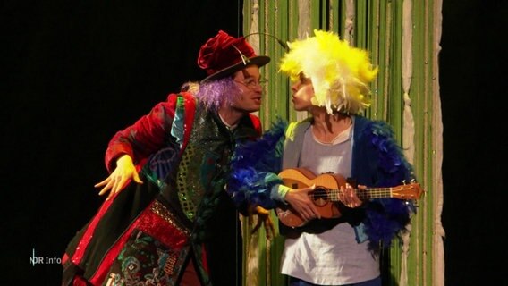 Eine Schauspielerin mit einer Gitarre unterhält sich mit einem Schauspieler auf einer Bühne. © Screenshot 