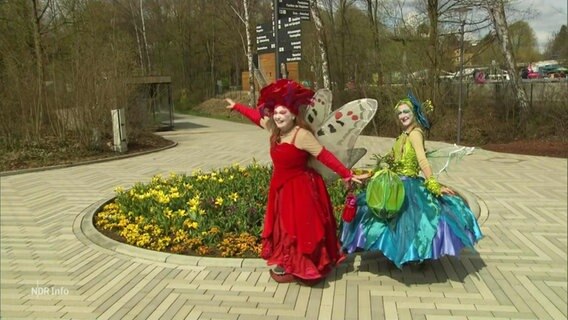 Zwei als Schmetterlinge verkleidete Schauspielerinnen stehen vor einem Blumenbeet. © Screenshot 