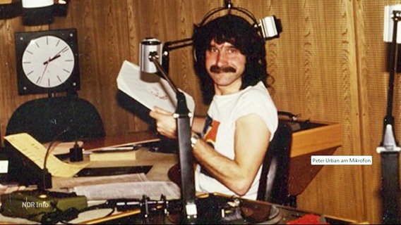 Peter Urban in jungen Jahren. Er sitzt in einem Studio, trägt lange Haare und einen Schnauzer. © Screenshot 