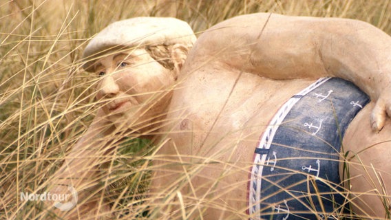 Eine lebensgroße Skulptur eines Mannes mit Schiebermütze und in Badehose liegt im Dünengras. © Screenshot 