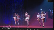 Mehrere Musicaldarstellende tanzen auf einer unt glitzernden Bühne im Scheinwerferlicht in Synchronität. © Screenshot 