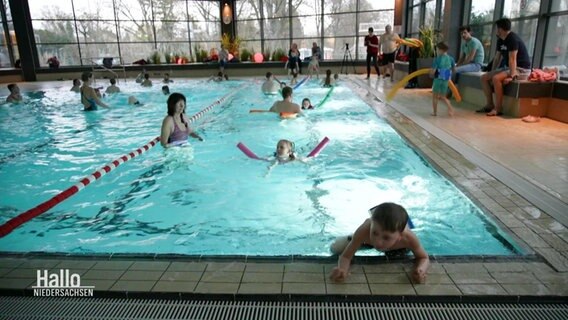 In einem Becken eines Schwimmbads schwimmen mehrere Kinder mit Schwimmnudeln ihre Bahnen. © Screenshot 