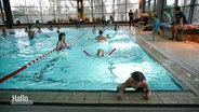 In einem Becken eines Schwimmbads schwimmen mehrere Kinder mit Schwimmnudeln ihre Bahnen. © Screenshot 