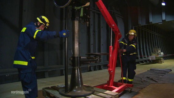 Zwei Einsatzkräfte des THW bewegen ein größeres Stahlstück mit einer Hebemaschine in einer Halle. © Screenshot 