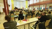 Mehrere Personen sitzen in einem größeren Sitzungssaal an mehreren Tischreihen. © Screenshot 