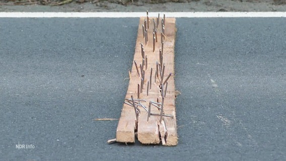 Auf Straßenasphalt liegt ein Holzbrett mit mehreren hineingeschlagenen Nägeln. © Screenshot 